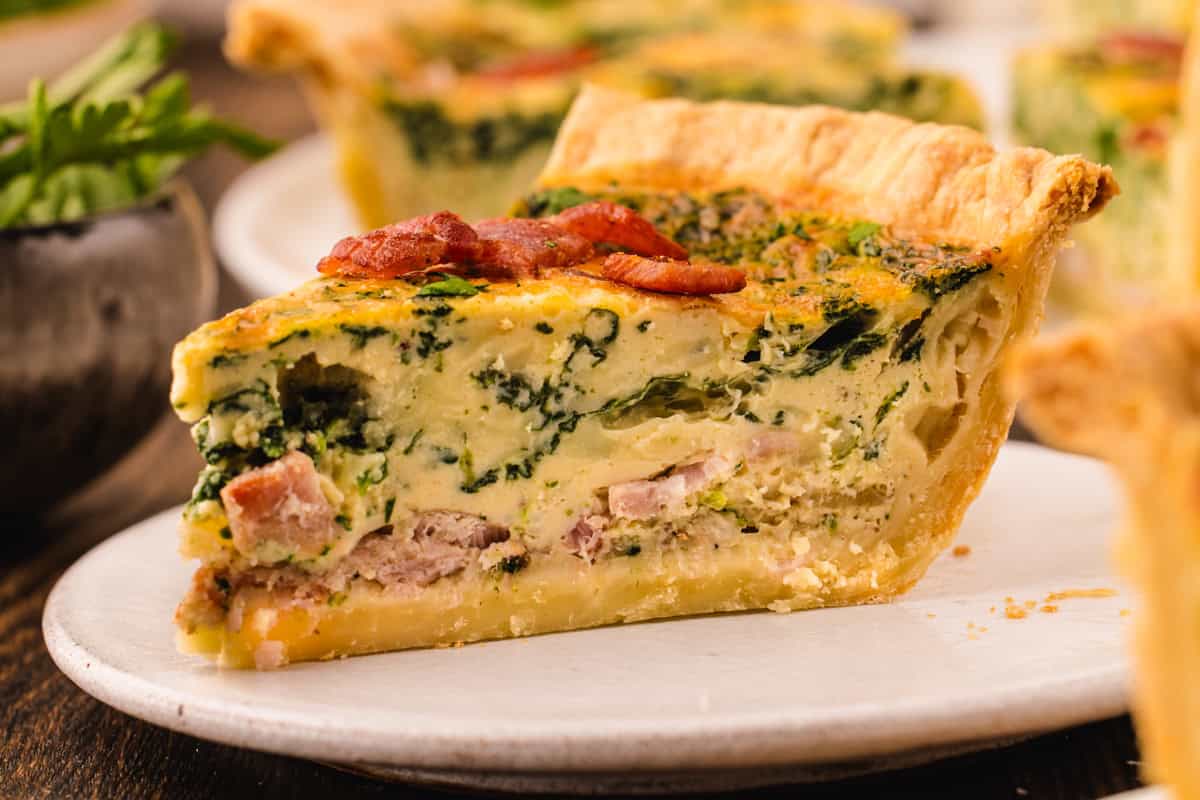 A slice of spinach and bacon quiche with a golden crust on a white plate.