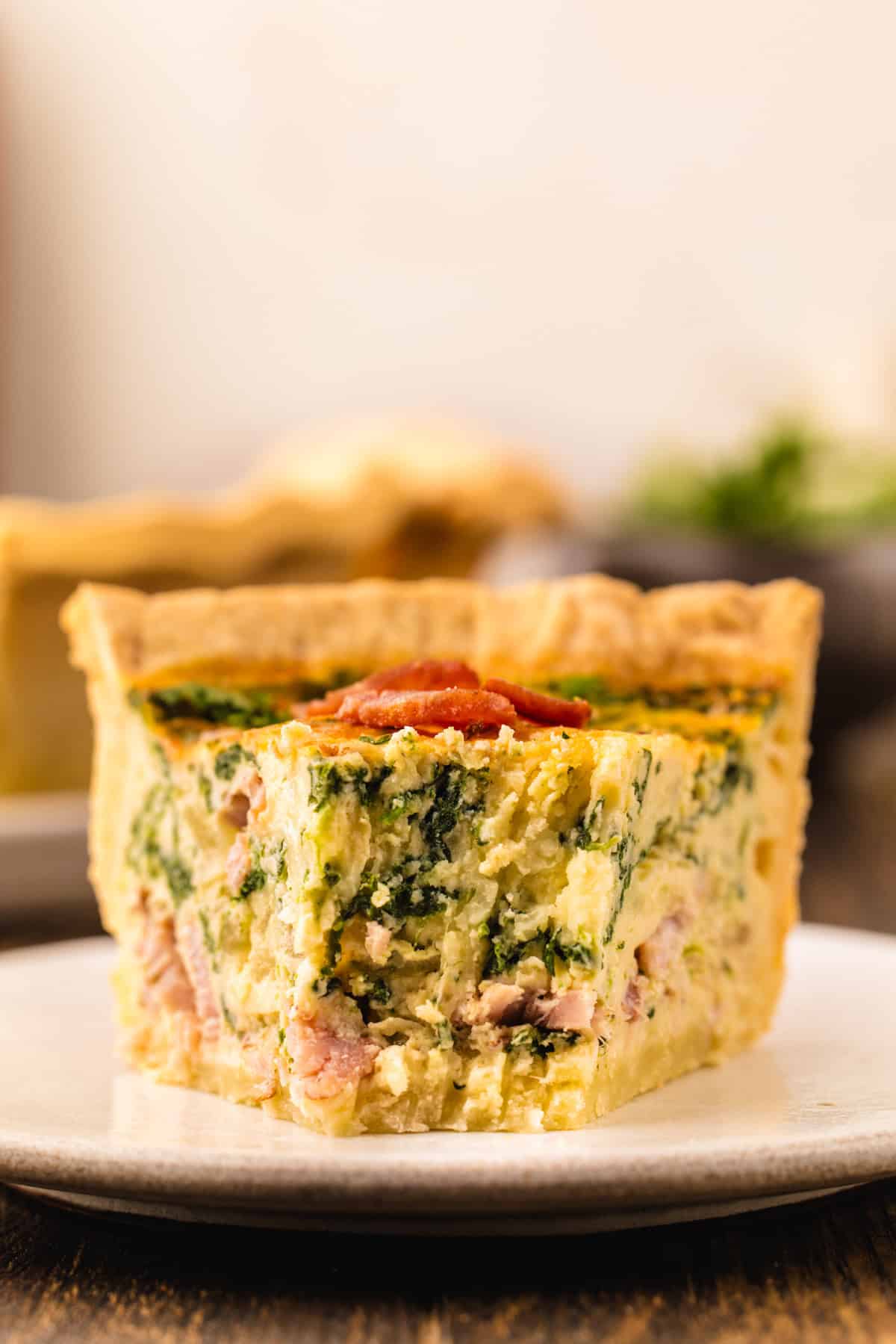 Quiche A single slice of quiche filled with spinach and bacon, topped with a piece of bacon on a small plate.