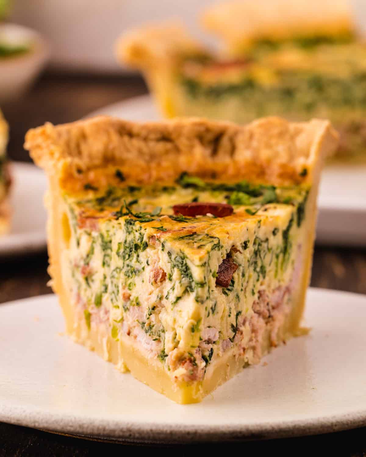Quiche A slice of quiche with spinach, bacon, and herbs in a flaky crust, served on a white plate.