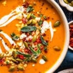 A bowl of orange soup garnished with swirls of cream, pumpkin seeds, fresh herbs, and crumbles of cheese.