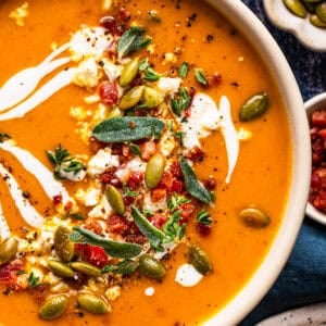 A bowl of orange soup garnished with swirls of cream, pumpkin seeds, fresh herbs, and crumbles of cheese.