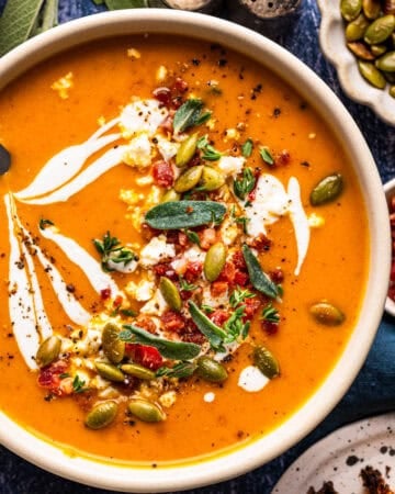 A bowl of orange soup topped with a swirl of cream, crumbled cheese, bacon, sage leaves, and pumpkin seeds.