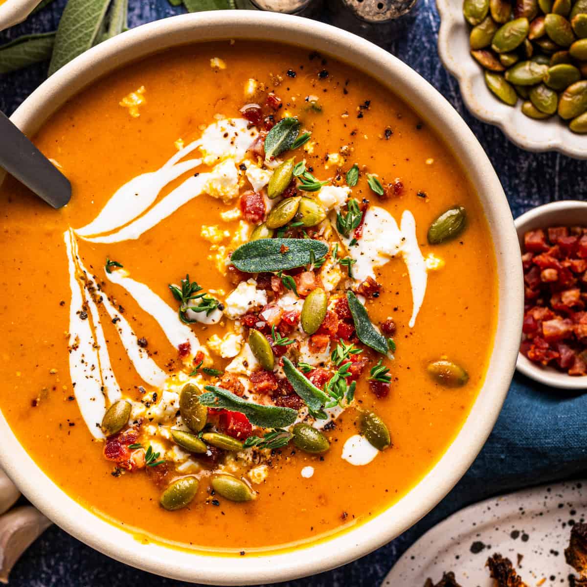 A bowl of orange soup topped with a swirl of cream, crumbled cheese, bacon, sage leaves, and pumpkin seeds.
