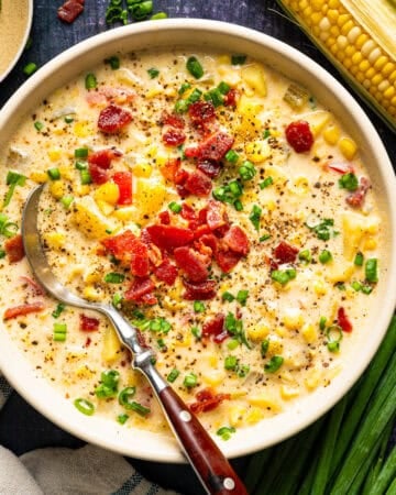 A bowl of creamy corn chowder topped with crumbled bacon and chopped green onions, with a silver spoon inside.