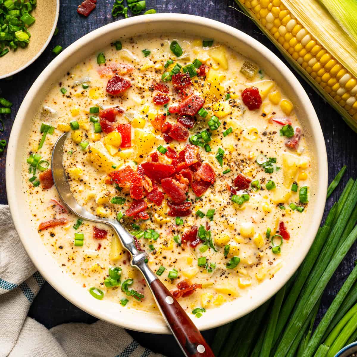 A bowl of creamy corn chowder topped with crumbled bacon and chopped green onions, with a silver spoon inside.