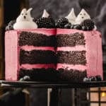 A three-layer chocolate cake with pink frosting, topped with blackberries, blueberries, and ghost-shaped meringues, with a slice removed.