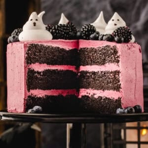 A three-layer chocolate cake with pink frosting, topped with blackberries, blueberries, and ghost-shaped meringues, with a slice removed.