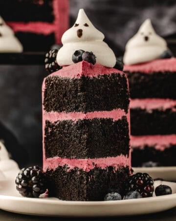 A slice of layered chocolate cake with pink frosting, topped with a ghost-shaped meringue, blueberries, and blackberries on a white plate.