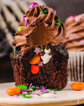 A chocolate cupcake with chocolate frosting and a hollowed-out center filled with Halloween-themed sprinkles, including a ghost, pumpkin, and star shapes.