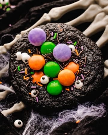 A dark chocolate cookie topped with colorful candy-coated chocolates, candy eyes, and chocolate sprinkles, resting on a skeleton hand prop decorated with faux spiderwebs.