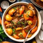A bowl of beef stew with potatoes and carrots, garnished with parsley and served with a slice of crusty bread.