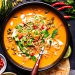 A bowl of orange soup garnished with fried shallots, pumpkin seeds, fresh herbs, and a drizzle of cream, served with a spoon and flatbread.