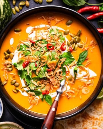 A bowl of orange soup topped with pumpkin seeds, fried shallots, herbs, chili slices, and a swirl of cream, served with a side of flatbread and lime.