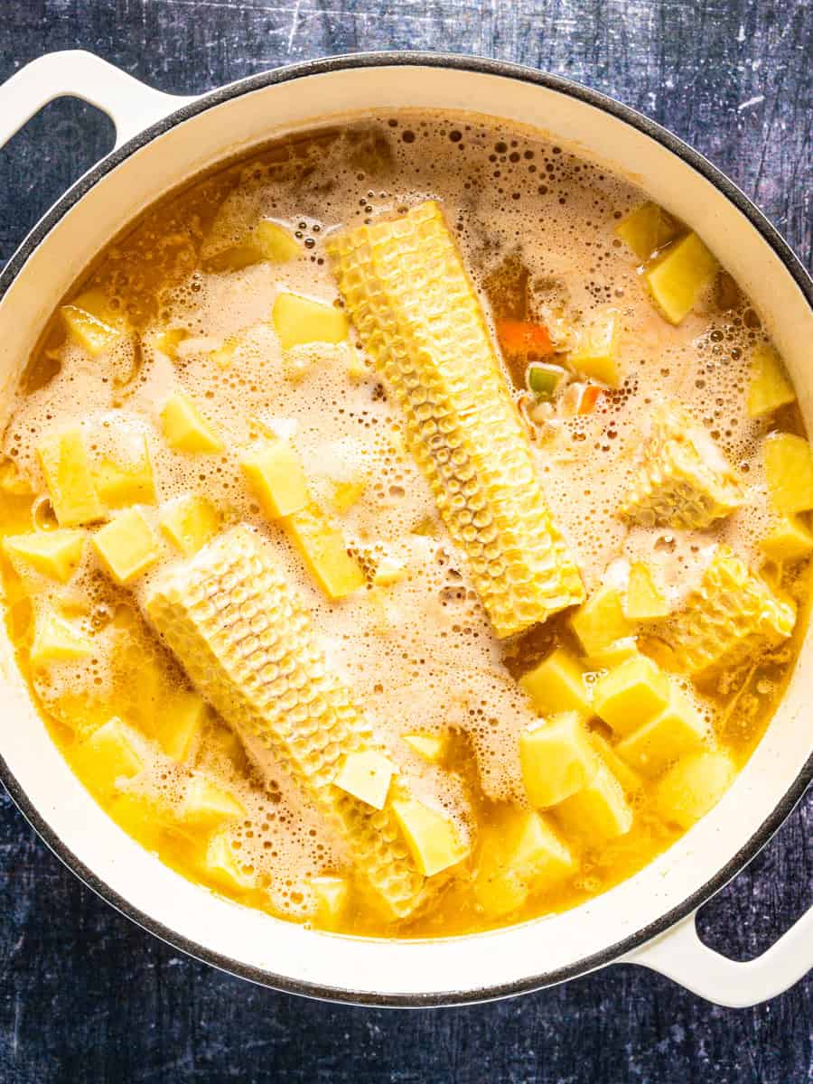 How to Make Corn Chowder (Step-By-Step Photos) A white pot containing boiling soup with corn cobs and diced potatoes.