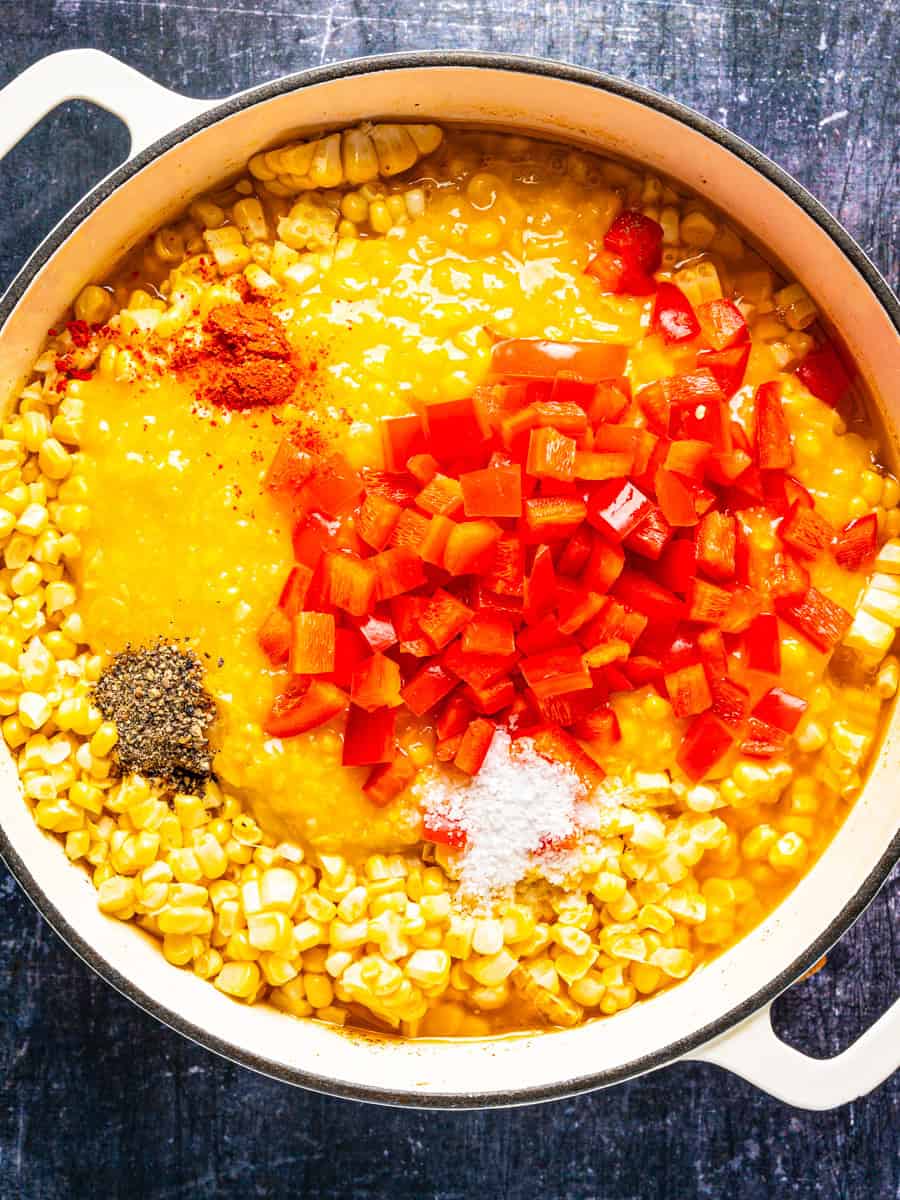How to Make Corn Chowder (Step-By-Step Photos) A white pot filled with corn kernels, a creamy yellow base, diced red bell peppers, paprika, black pepper, and salt.