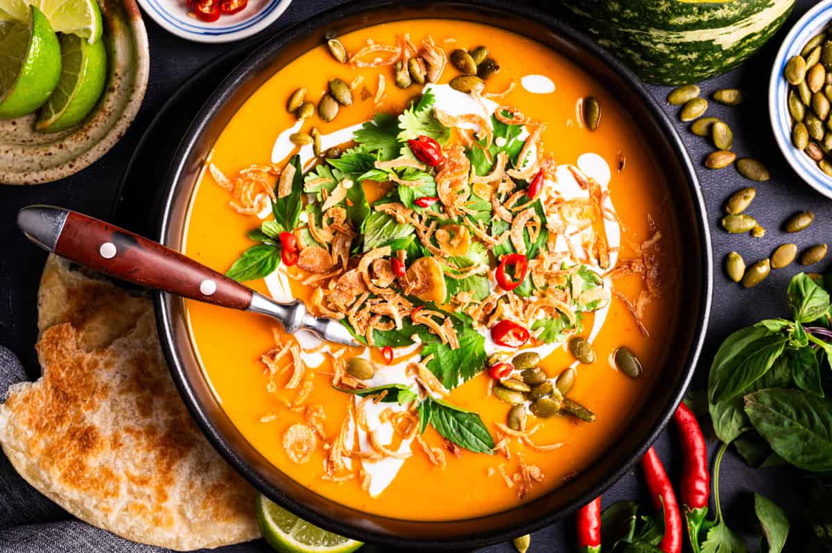 A bowl of creamy orange soup topped with swirls of cream, fresh herbs, crispy fried onions, and pumpkin seeds.