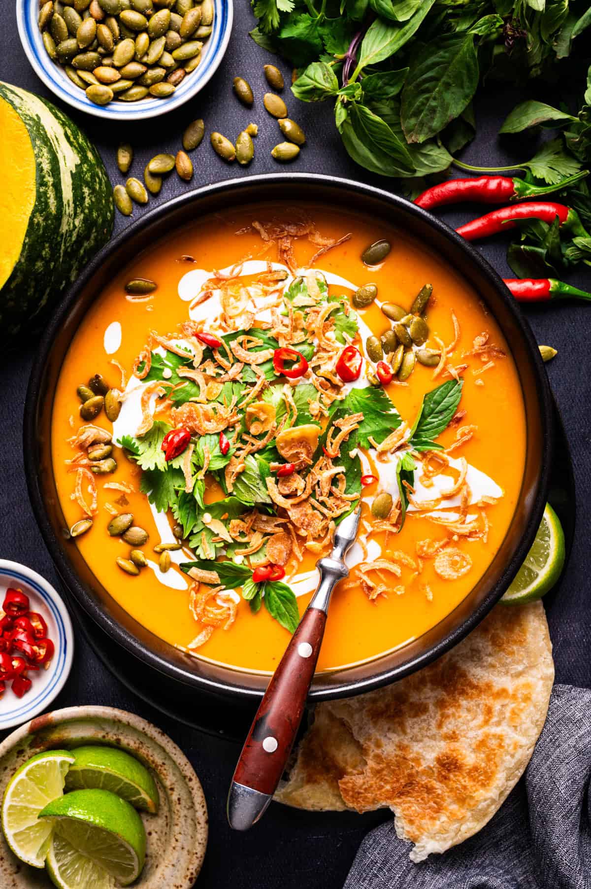Thai Pumpkin Soup A bowl of orange soup topped with herbs, fried shallots, chili slices, and seeds.