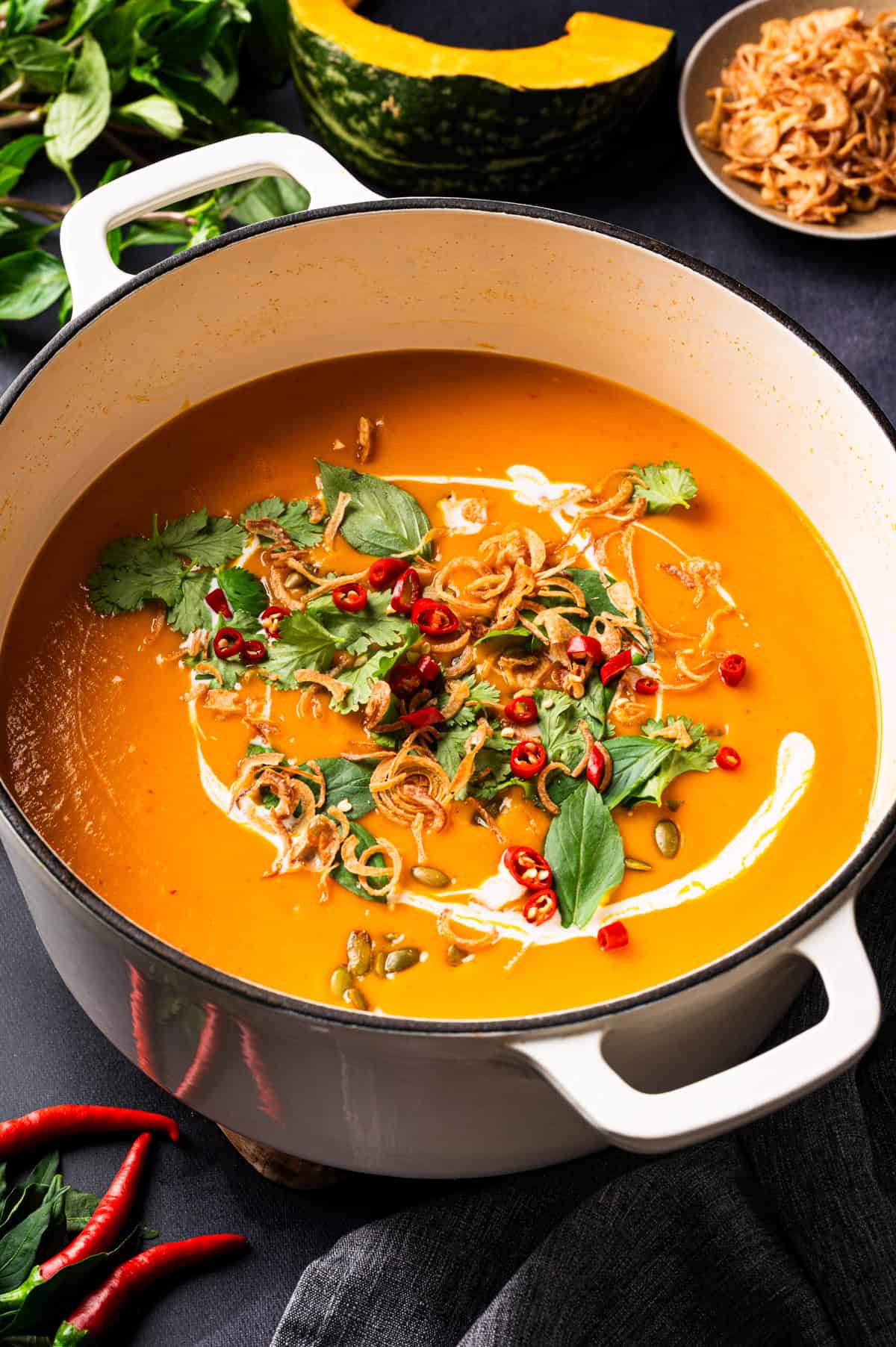 Thai Pumpkin Soup A large white pot filled with creamy orange soup, topped with swirls of cream, fresh cilantro, fried shallots, pumpkin seeds, and sliced red chili peppers.