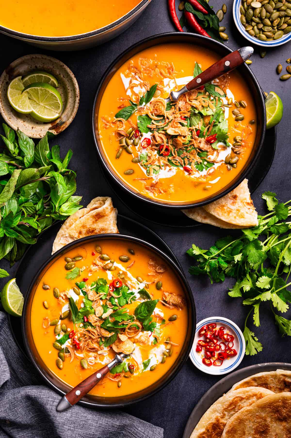 Thai Pumpkin Soup Two bowls of creamy orange soup topped with herbs, fried shallots, chili slices, and pumpkin seeds.