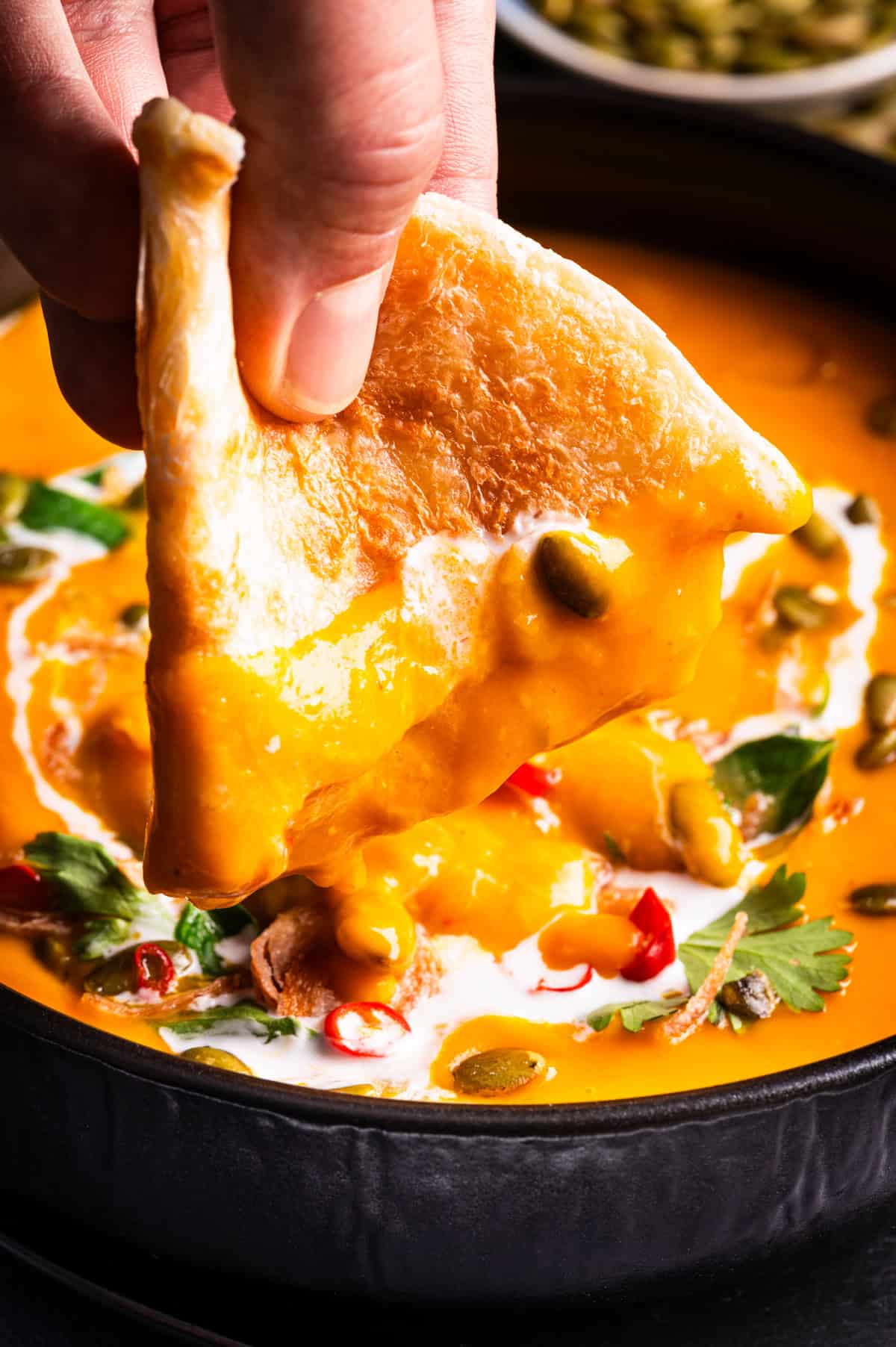 Thai Pumpkin Soup A hand dips a piece of flatbread into a bowl of creamy orange soup topped with herbs, chili slices, and seeds.