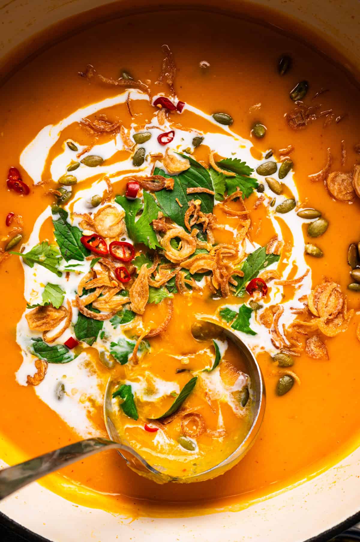 Thai Pumpkin Soup A metal ladle sits in a pot of orange soup garnished with a swirl of cream, fried shallots, pumpkin seeds, fresh herbs, and sliced red chilies.