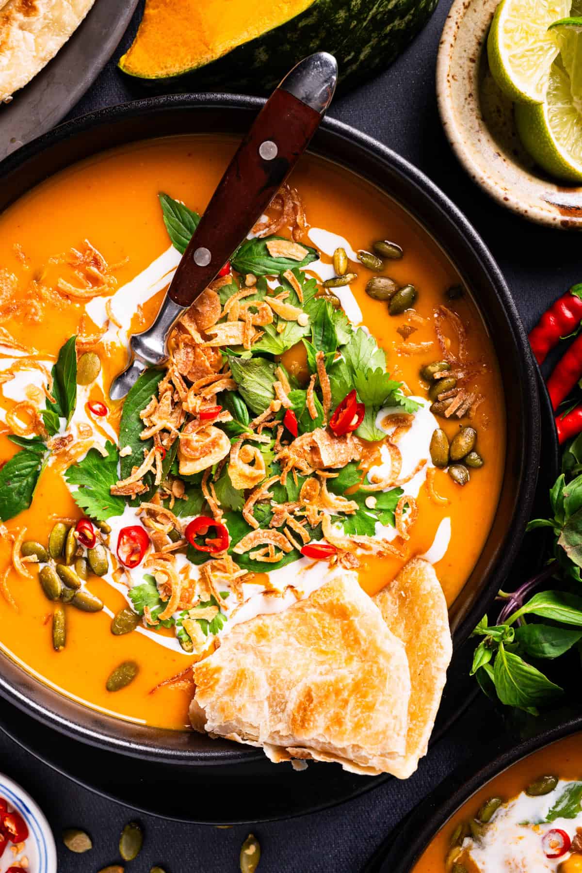 Thai Pumpkin Soup A bowl of orange soup garnished with fried shallots, pumpkin seeds, fresh herbs, chili slices, and a drizzle of cream, served with a piece of flatbread and a spoon.