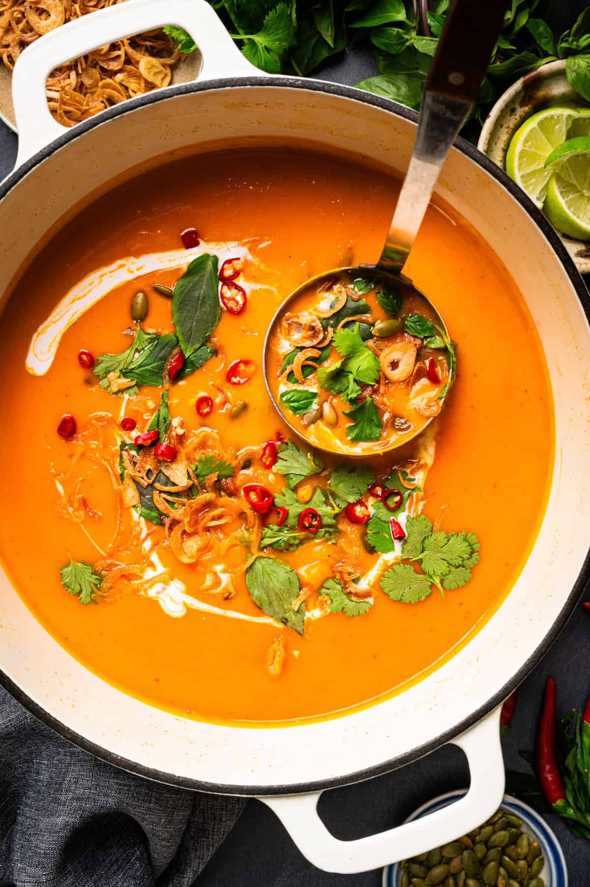 Thai Pumpkin Soup Creamy orange soup in a white pot, garnished with herbs, fried shallots, chili slices, and a swirl of cream, with a ladle resting inside.