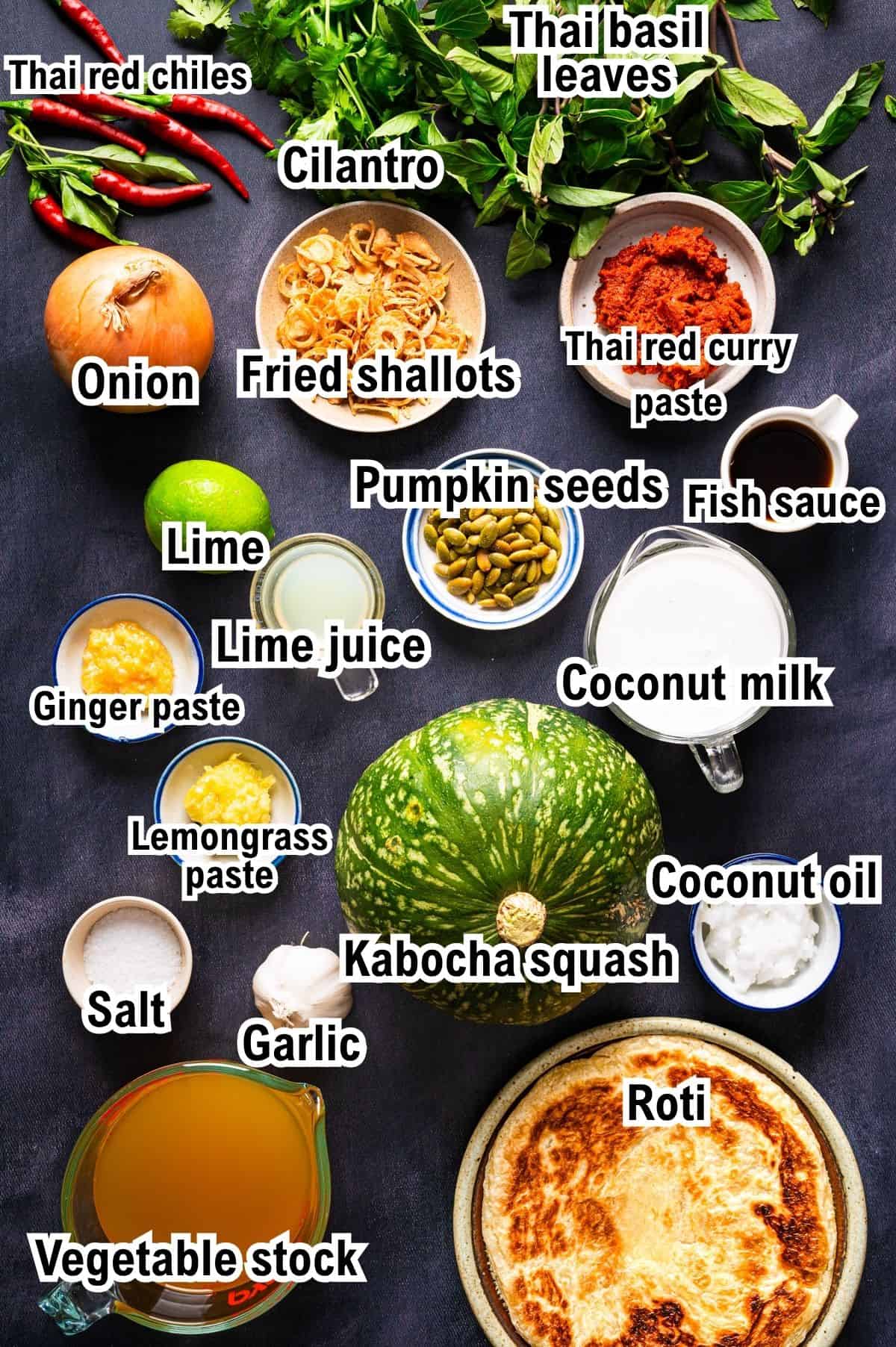 The Ingredients for Thai Pumpkin Soup This photo shows the ingredients for Thai Pumpkin Soup: a whole kabocha squash, roti, coconut milk, curry paste, aromatics, and various seasonings arranged on a dark surface.